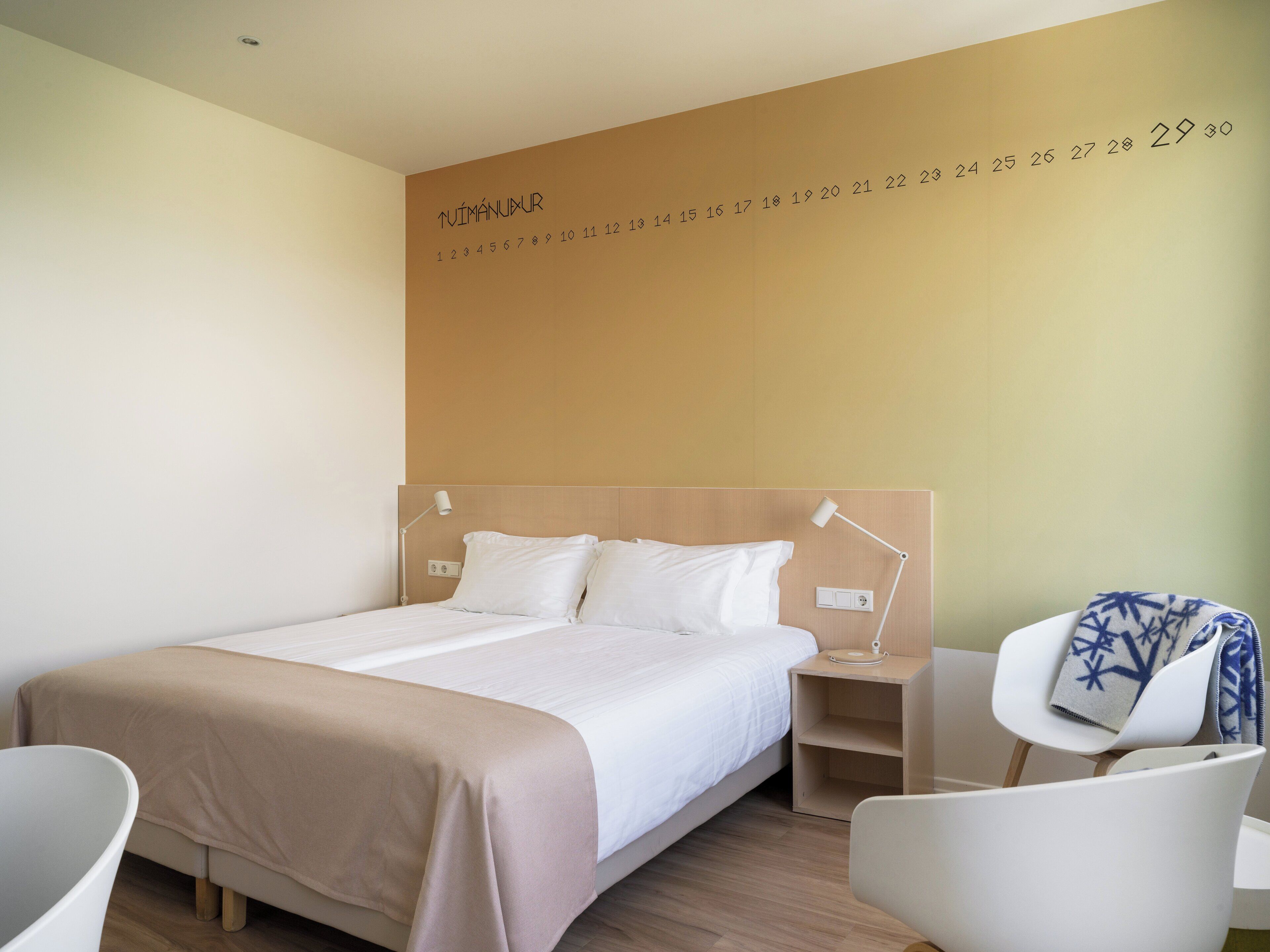 A hotel room with a bed, bedside tables, a chair, and a wall with numbers.