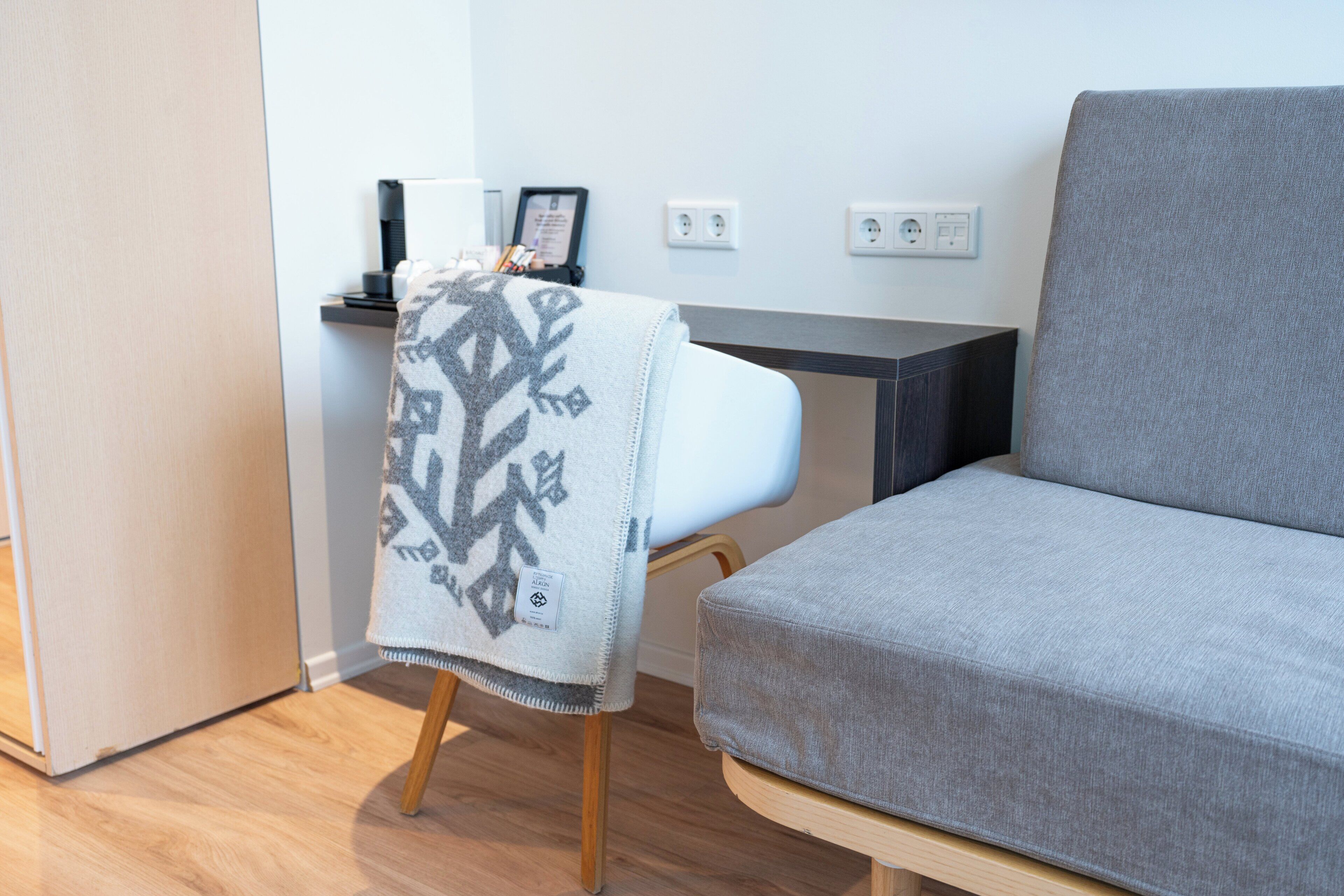 Una sala de estar moderna con un sofá gris, un sillón blanco con una manta estampada, piso de madera y una pequeña mesa con una lámpara y un marco de fotos.