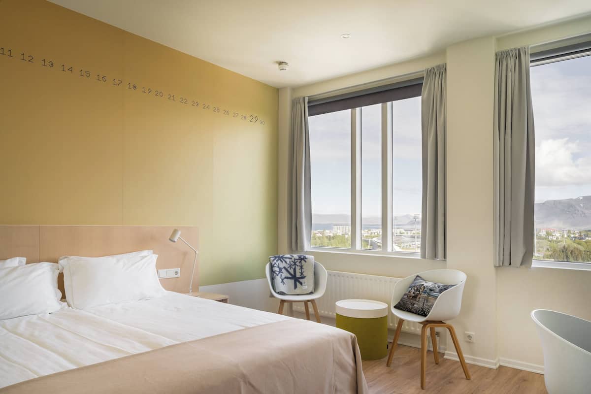 A hotel room with a bed, a chair, a small table, and a window with a city view.
