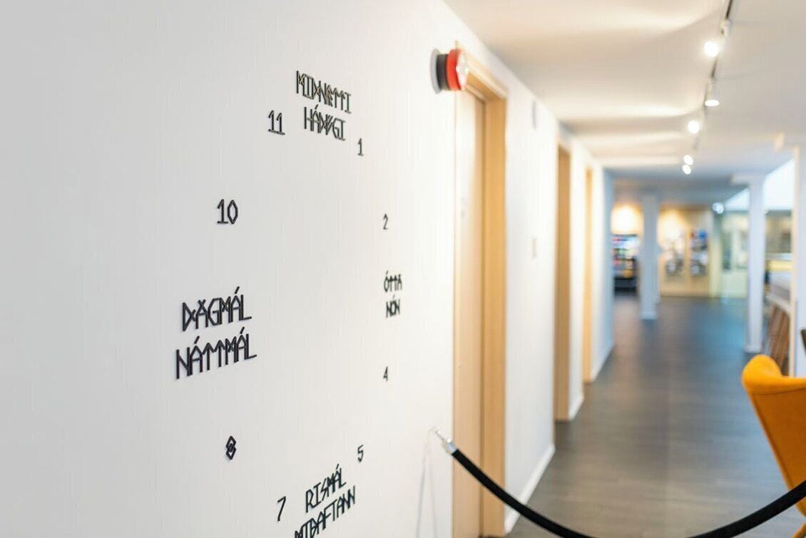 A corridor with a white wall displaying numbered labels and text, a fire alarm, and a yellow chair.
