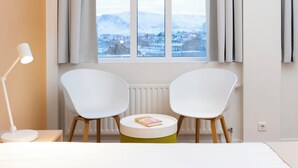 A room with two white chairs, a small round table, and a lamp. A book is on the table. The room has a window with a view of a snowy landscape and buildings. - Reykjavik Lights by Keahotels (Reykjavik)