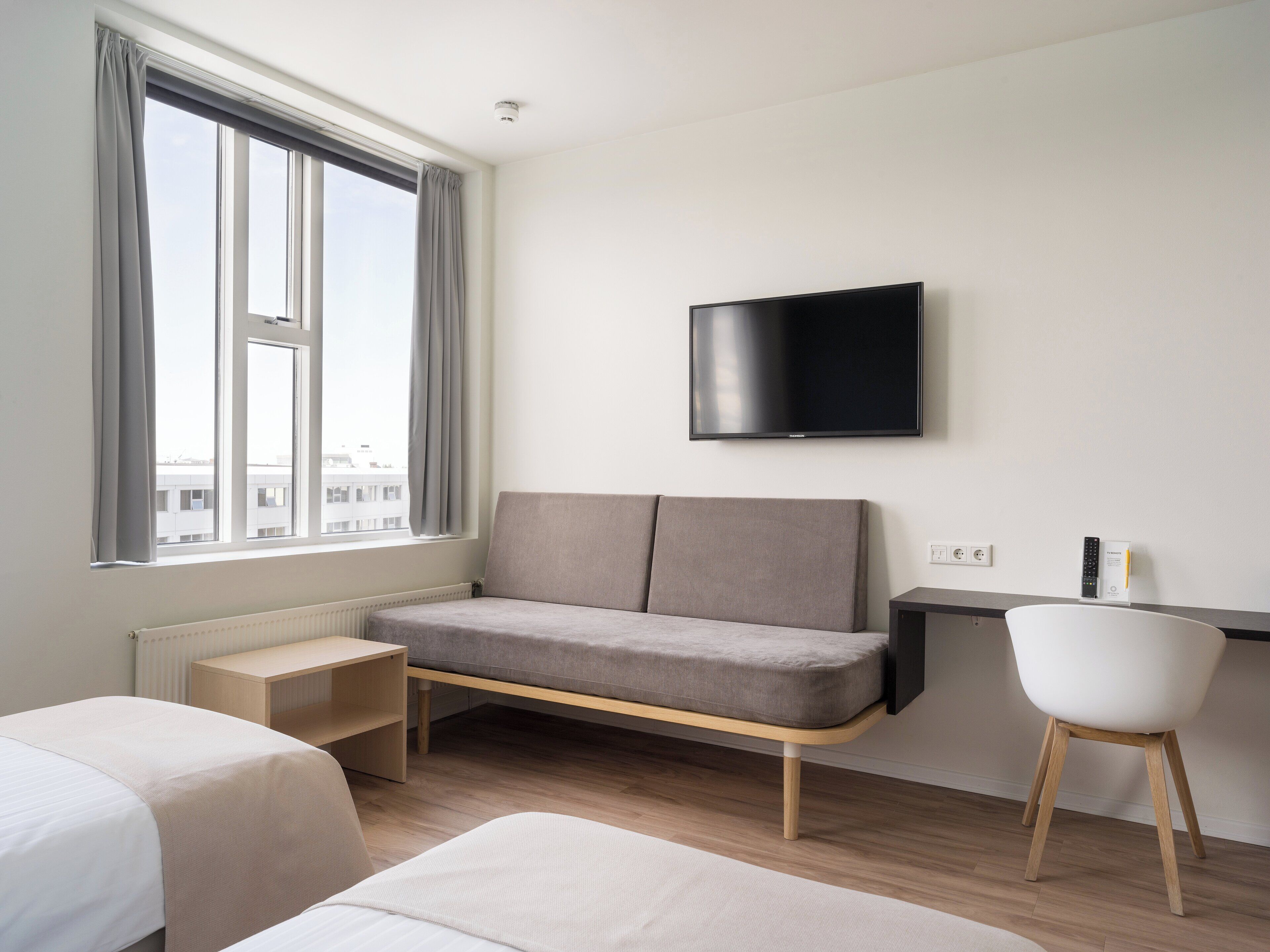 A modern hotel room with a large window, a flat-screen TV, a sofa, a desk, and a chair.