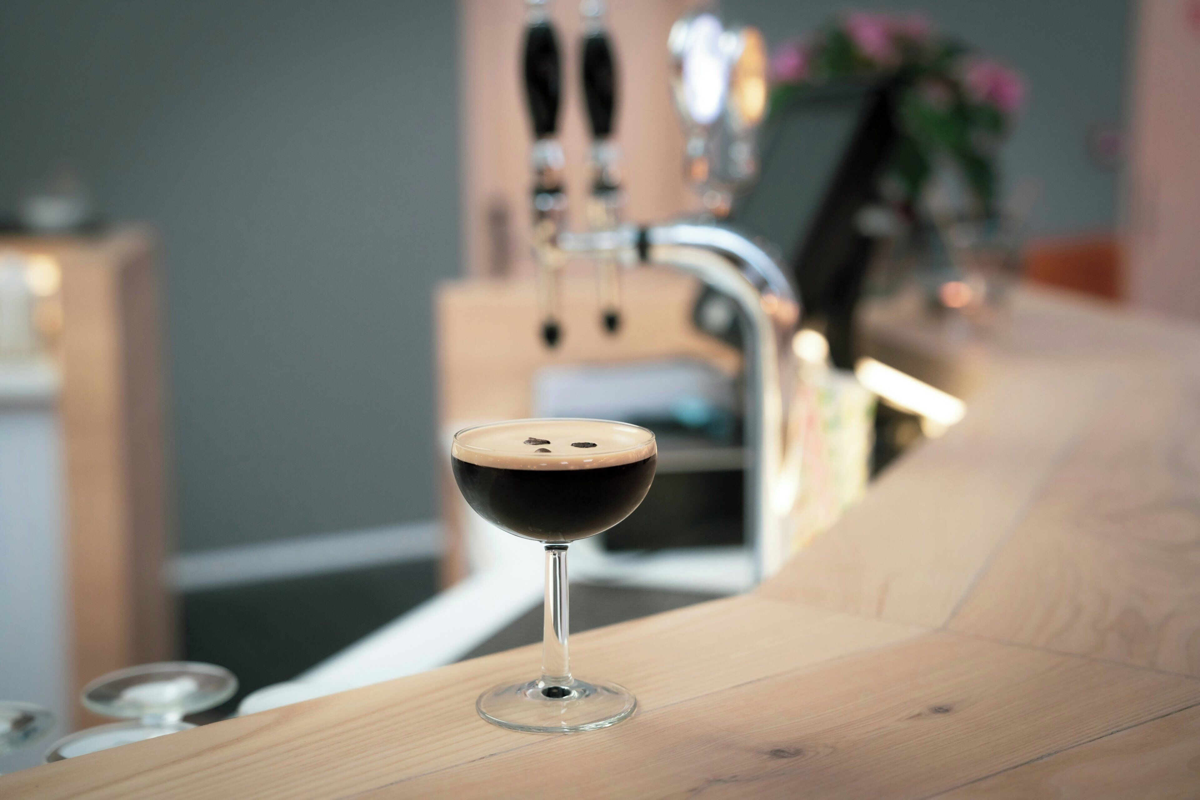 A bar setting with a glass of dark beverage, a bar counter, and a blurred background with a plant.