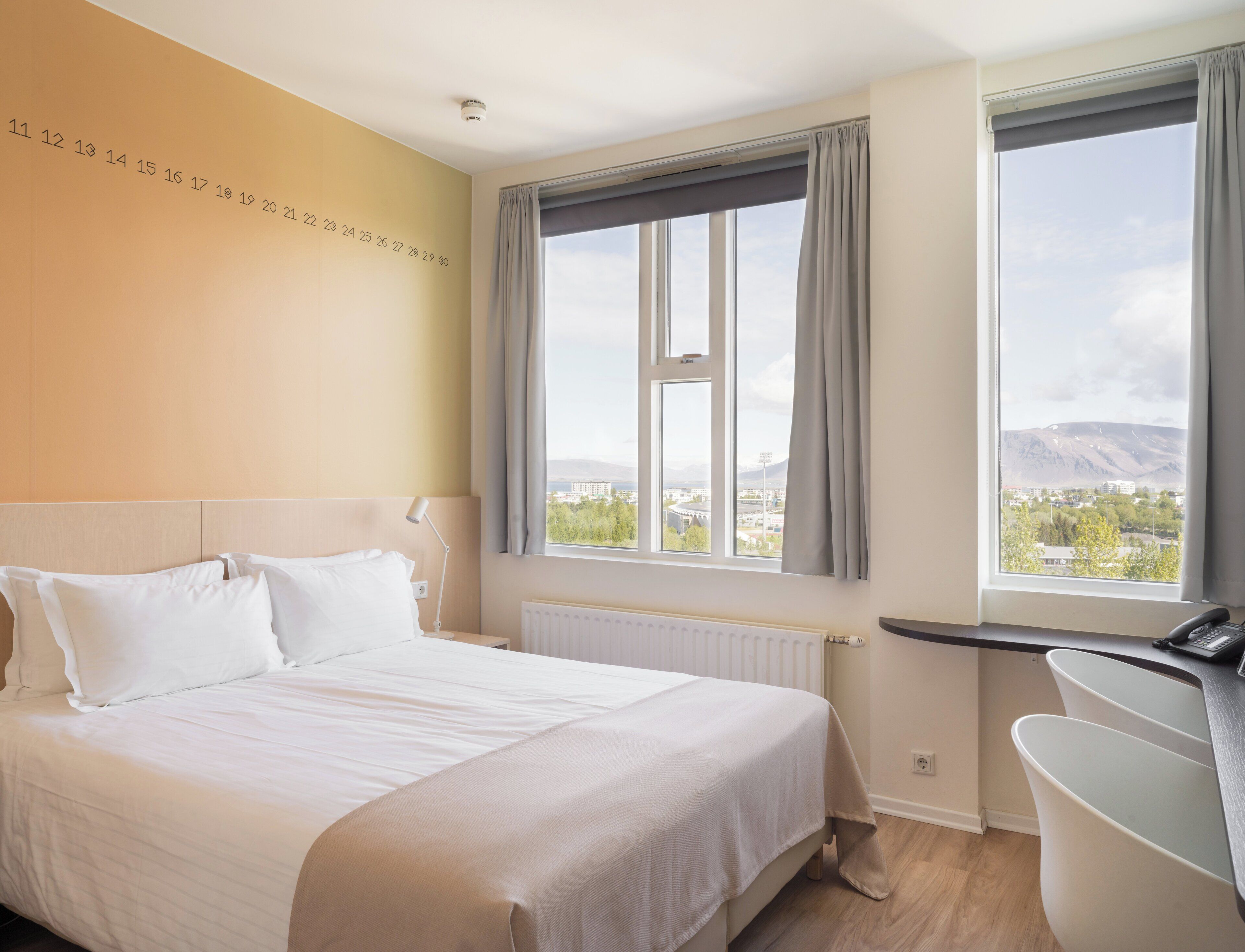 A hotel room with a bed, a desk, a view of a cityscape, and a wall with numbers.
