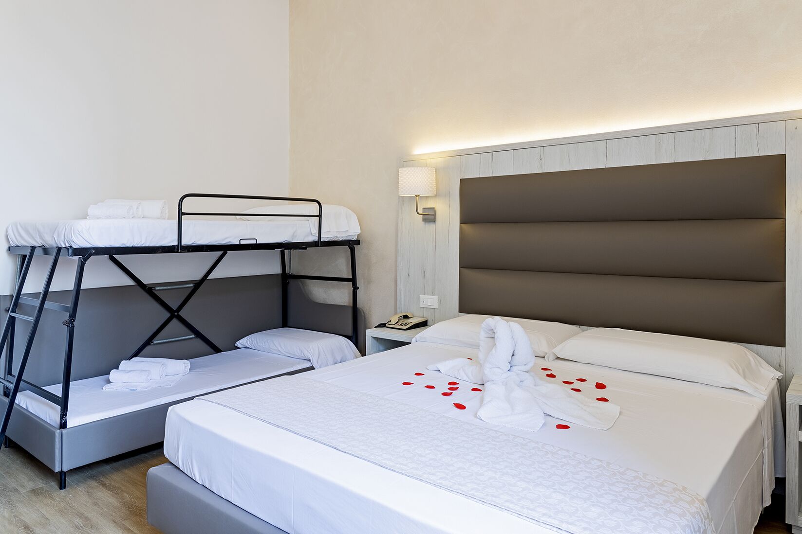 Quarto de hotel com uma cama beliche, uma cama de solteiro e uma mesa de cabeceira com abajur.