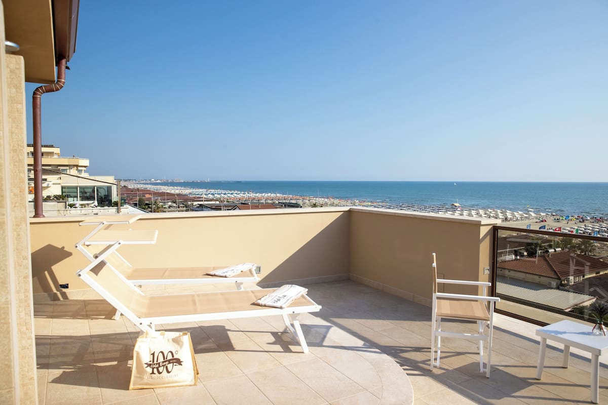 A balcony with lounge chairs and a table, overlooking a beach with umbrellas and a clear sky.