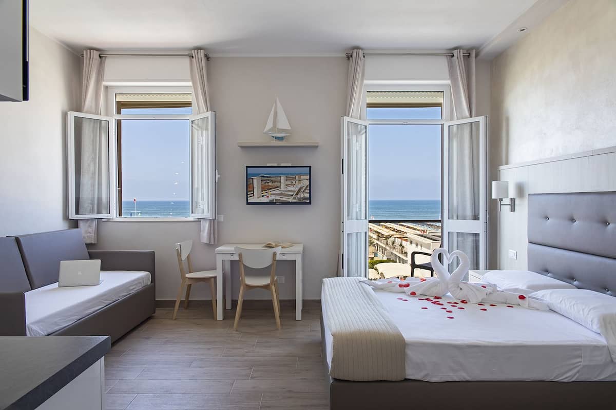 A hotel room with a bed, a desk, a chair, and two windows offering a view of the beach.