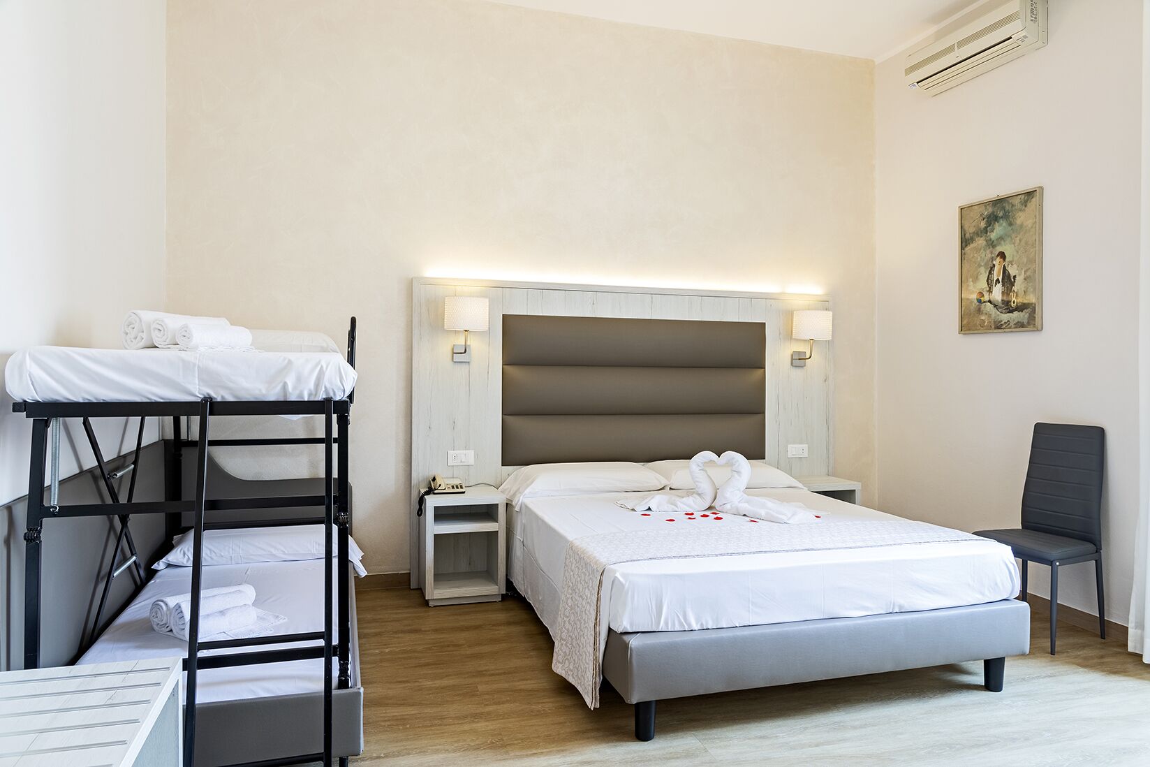 Een hotelkamer met een stapelbed, een eenpersoonsbed, nachtkastjes, een lamp, een stoel en een schilderij aan de muur.
