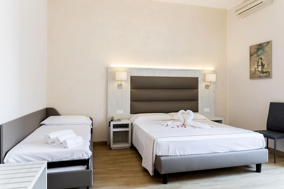 A hotel room with two single beds, a large bed, a bedside table with a lamp, a chair, and a painting on the wall.