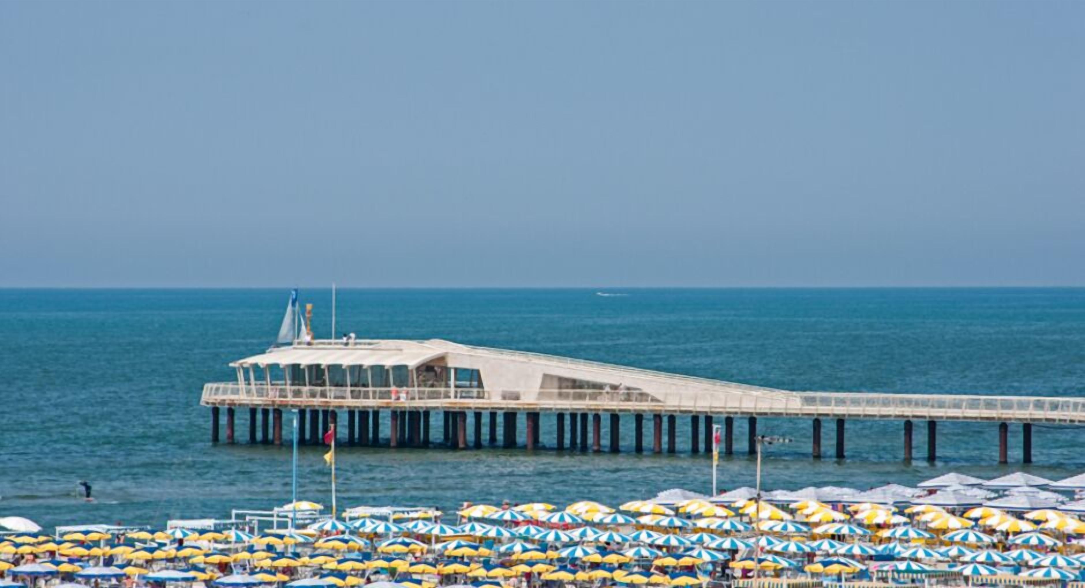 Un muelle que se adentra en el mar con un edificio en la parte superior, rodeado de numerosos sombrillas de playa.