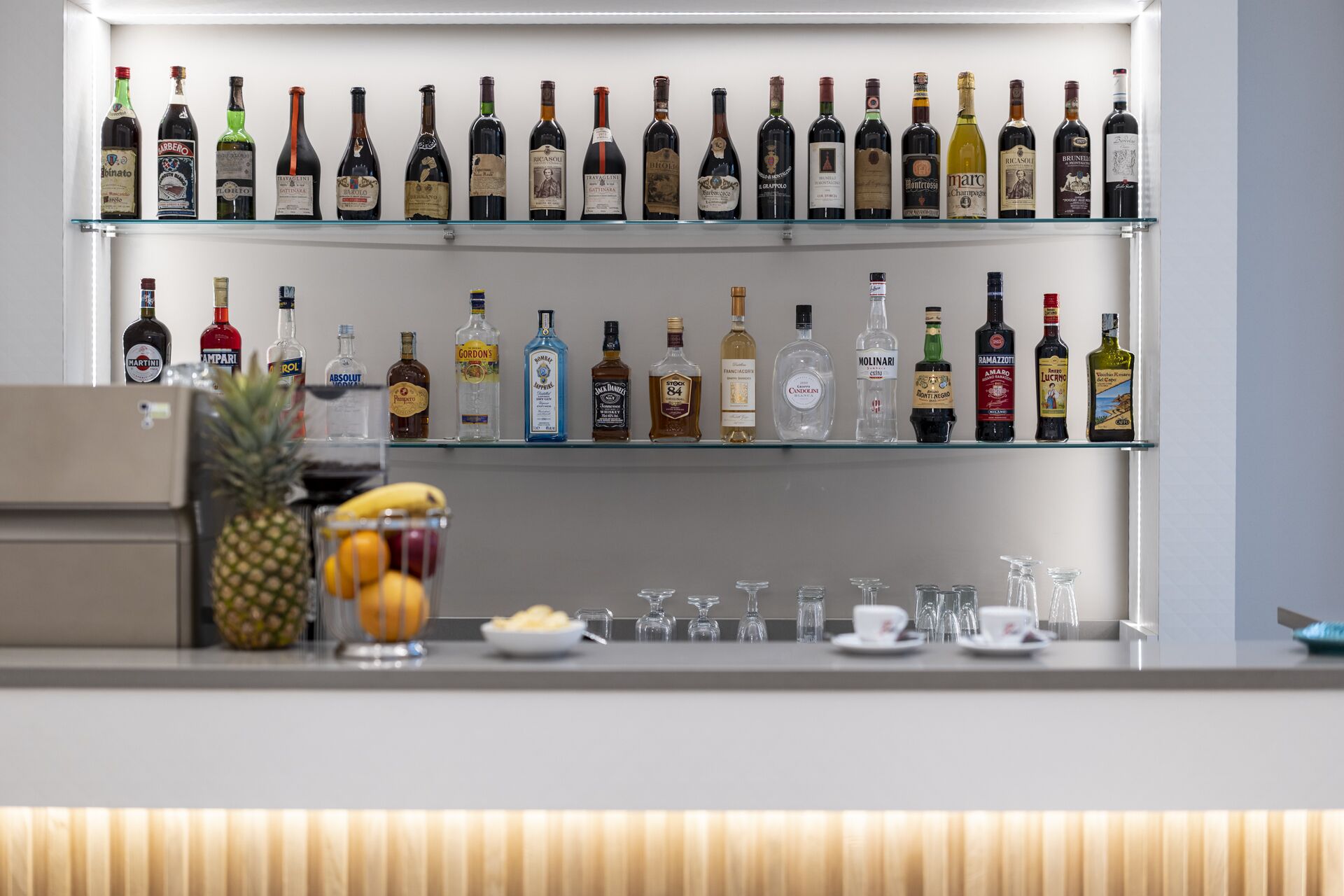 Zona de bar con varias estanterías llenas de botellas de licor, un frutero con piña y naranjas, y una variedad de vasos y copas.