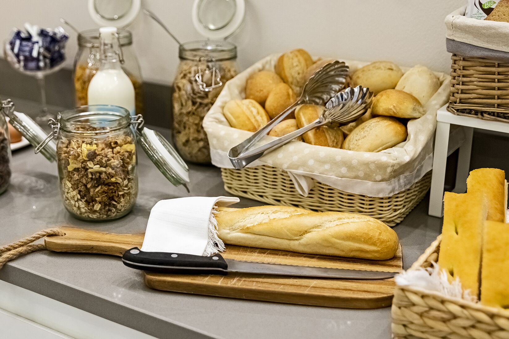A kitchen counter with various bread items, a loaf of bread, a knife, and a cutting board.
