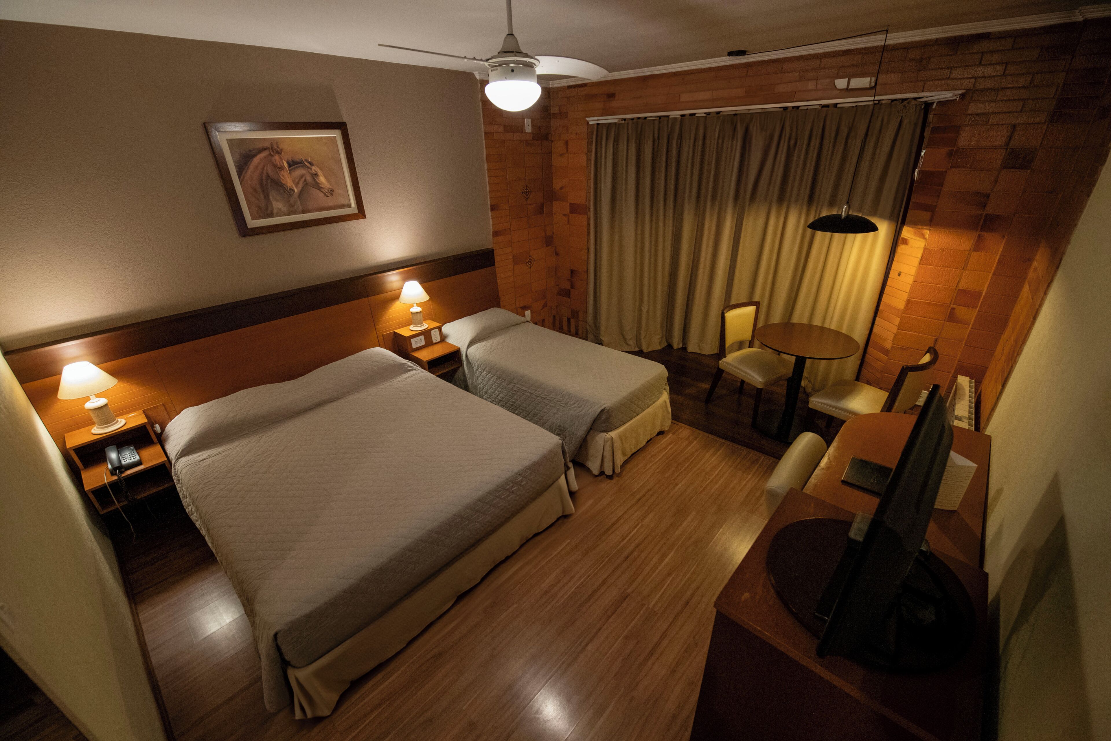 Een hotelkamer met twee bedden, een bureau, een stoel, een kleine tafel, een plafondventilator en een aan de muur hangend schilderij.