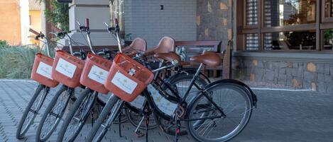 A row of bicycles with baskets, parked in front of a building with a brick facade and large windows.