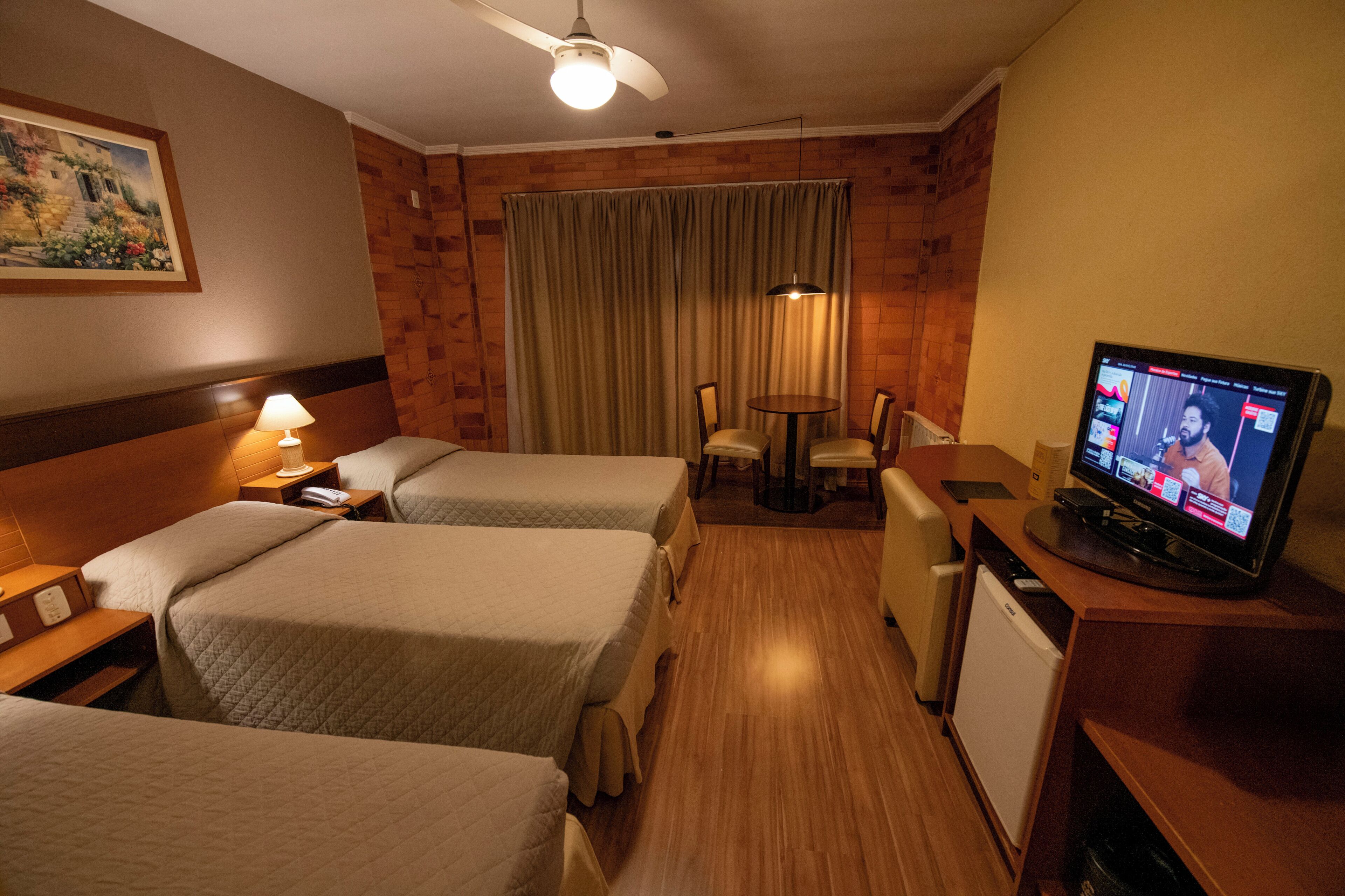 Habitación de hotel con dos camas, un escritorio, televisor, refrigerador pequeño y ventilador de techo.
