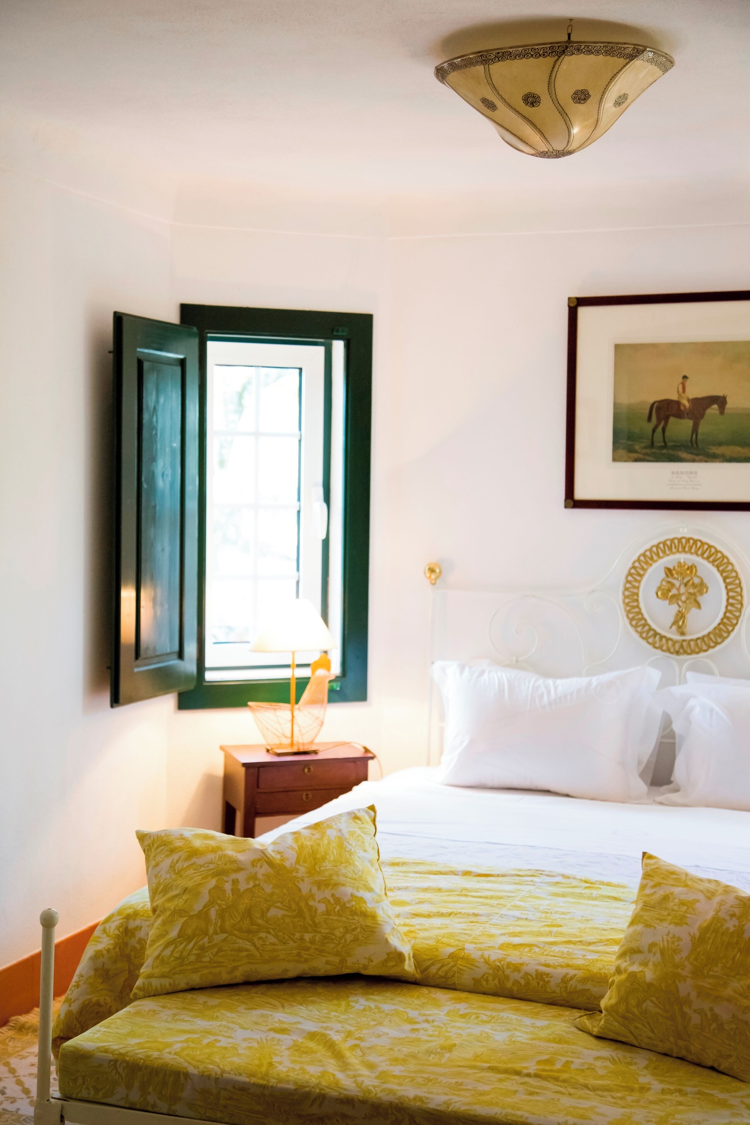 A room with a bed, a nightstand, a window with open shutters, and a framed picture on the wall.