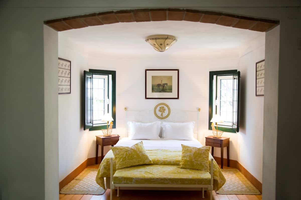 A bedroom with a bed, two nightstands, a framed picture on the wall, and two windows with open shutters.