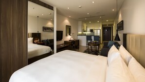 A hotel room with a large bed, a desk, a chair, a small table, a sofa, a TV, and a kitchen area in the background.