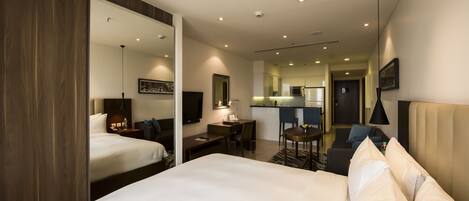 A hotel room with a large bed, a desk, a chair, a small table, a sofa, a TV, and a kitchen area in the background.