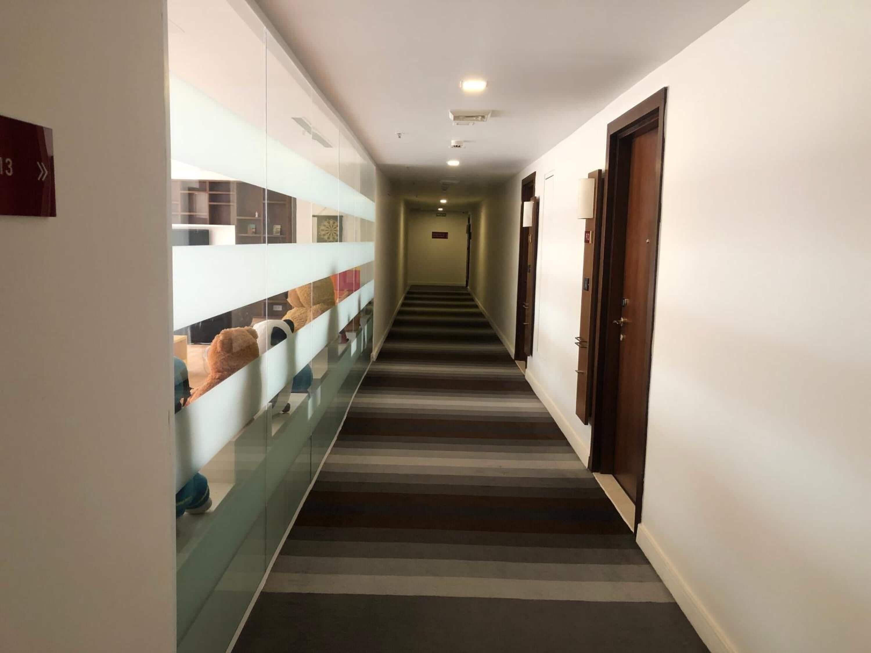 Passadís d'hotel amb un sòl de moqueta a ratlles, parets de vidre amb objectes decoratius i portes de fusta.