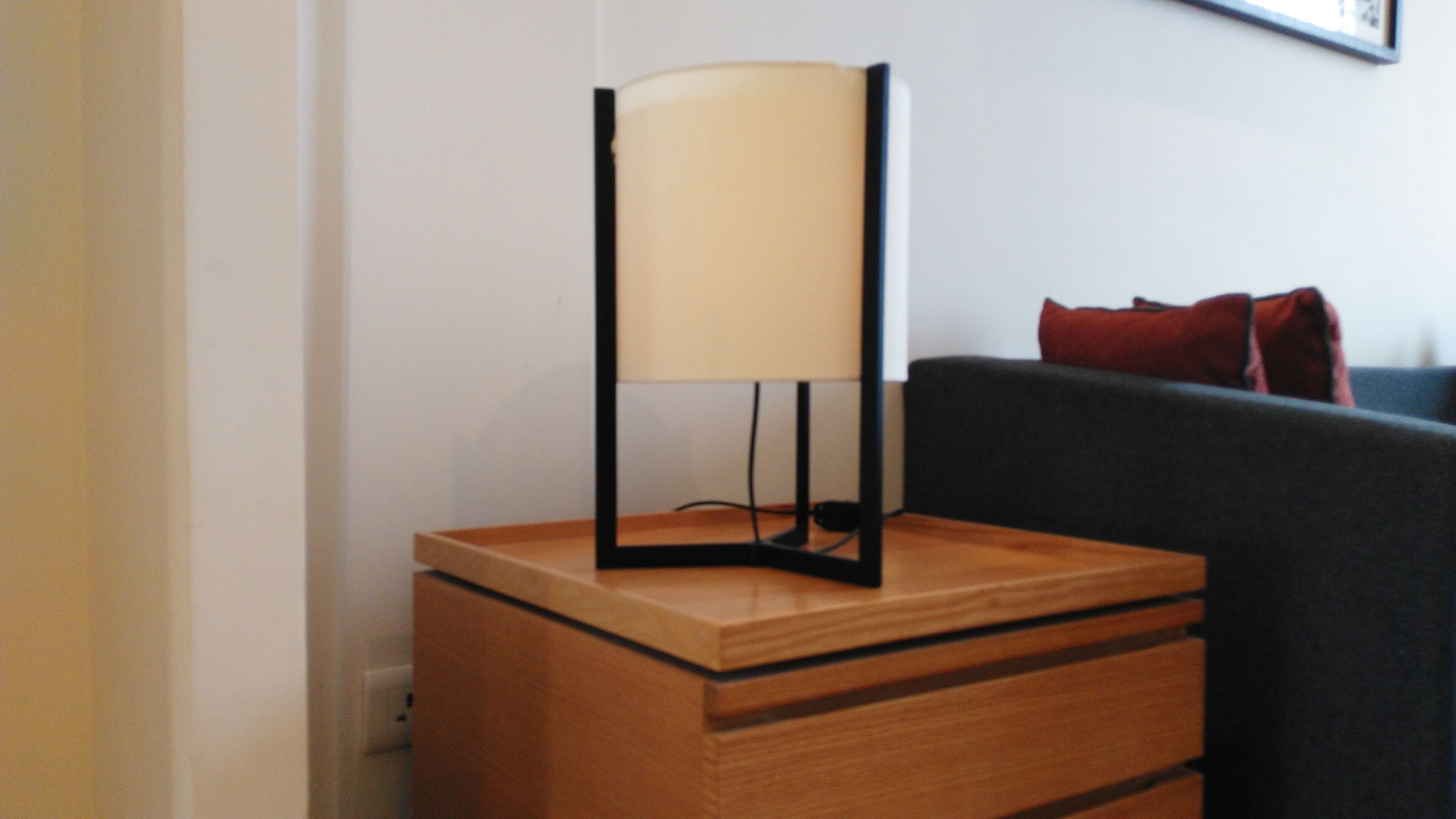 A wooden bedside table with a lamp and a drawer, a dark sofa with red cushions, and a white wall with a framed picture.
