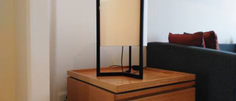 A wooden bedside table with a lamp and a drawer, a dark sofa with red cushions, and a white wall with a framed picture.