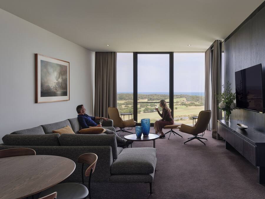 A modern hotel room with a large window, a flat-screen TV, a sofa, a dining table, and a coffee table.
