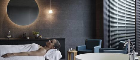 Une salle de bain moderne avec une grande baignoire, un miroir rond et un coin salon comprenant une chaise et une petite table.