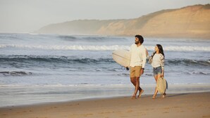 Ein Paar spaziert am Strand, ein Surfbrett tragend, mit Wellen im Meer und einer hügeligen Landschaft im Hintergrund.