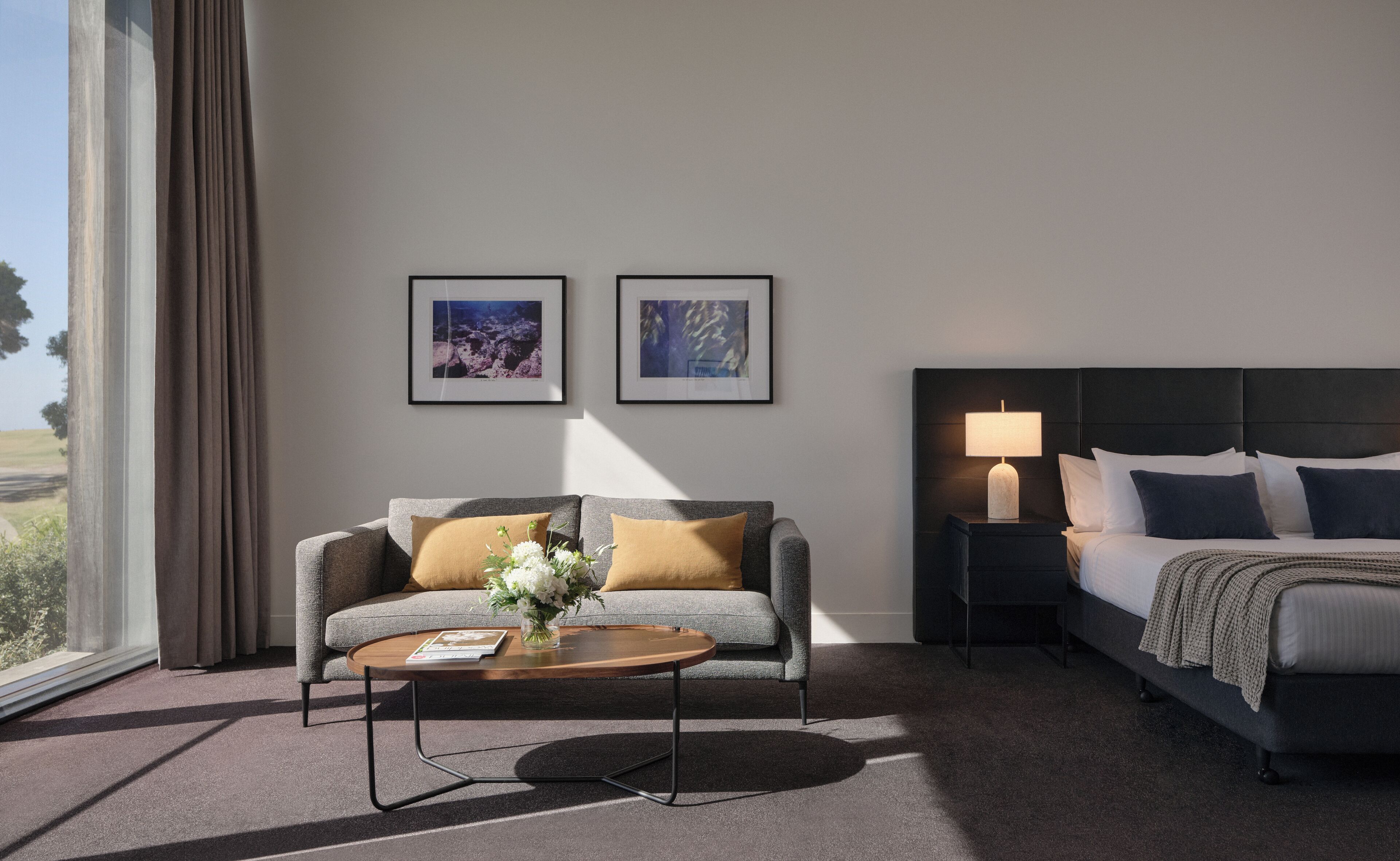 A hotel room with a sofa, a bed, a nightstand with a lamp, and two framed pictures on the wall.