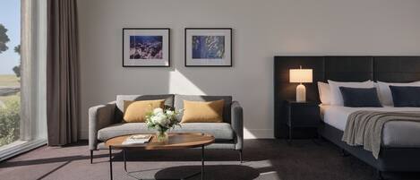 A hotel room with a sofa, a bed, a nightstand with a lamp, and two framed pictures on the wall.