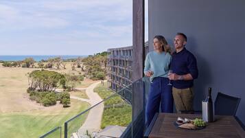 A balcony with a view of a golf course and ocean, a table with a bottle and grapes, and a person holding a glass.
