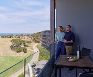 A balcony with a view of a golf course and ocean, a table with a bottle and grapes, and a person holding a glass.