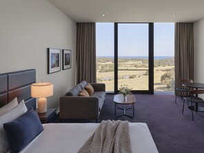 A hotel room with a bed, a sofa, a small table with a vase, and a desk with chairs. The room has large windows with a view of a landscape.