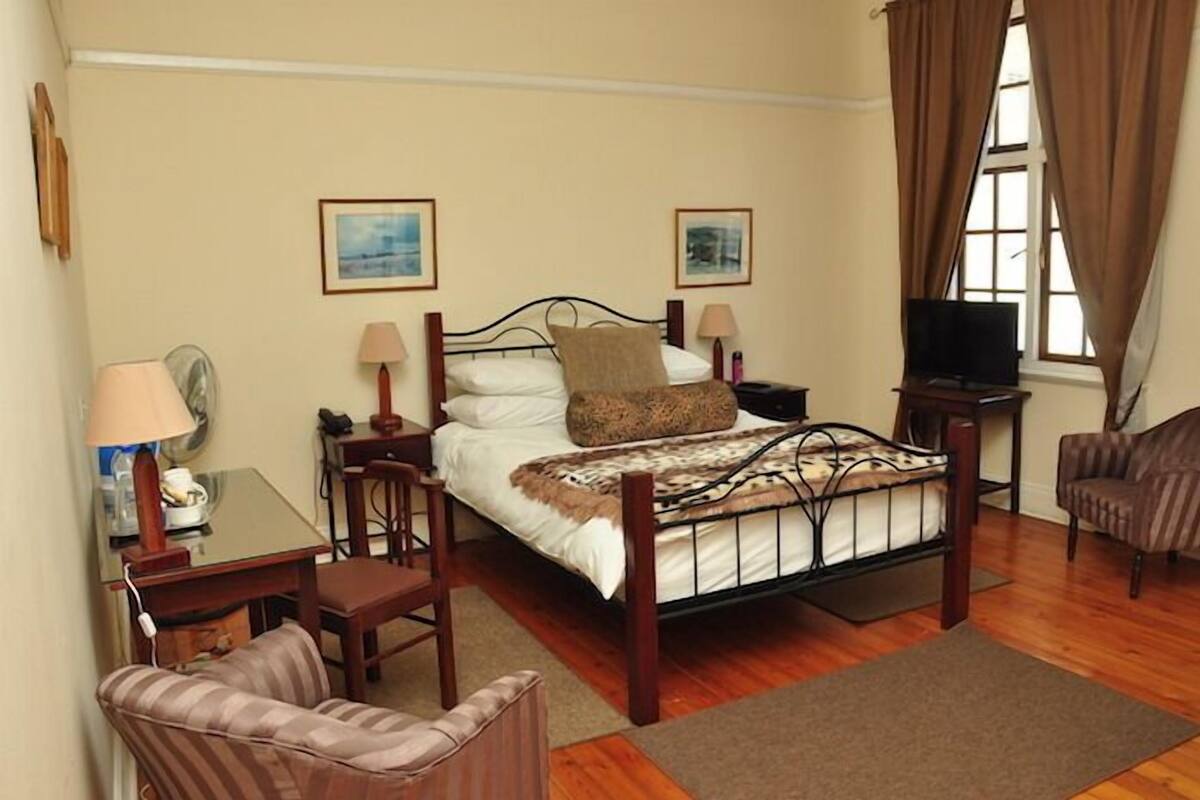 A hotel room with a bed, a TV, a desk with a lamp, a chair, and a window with curtains.