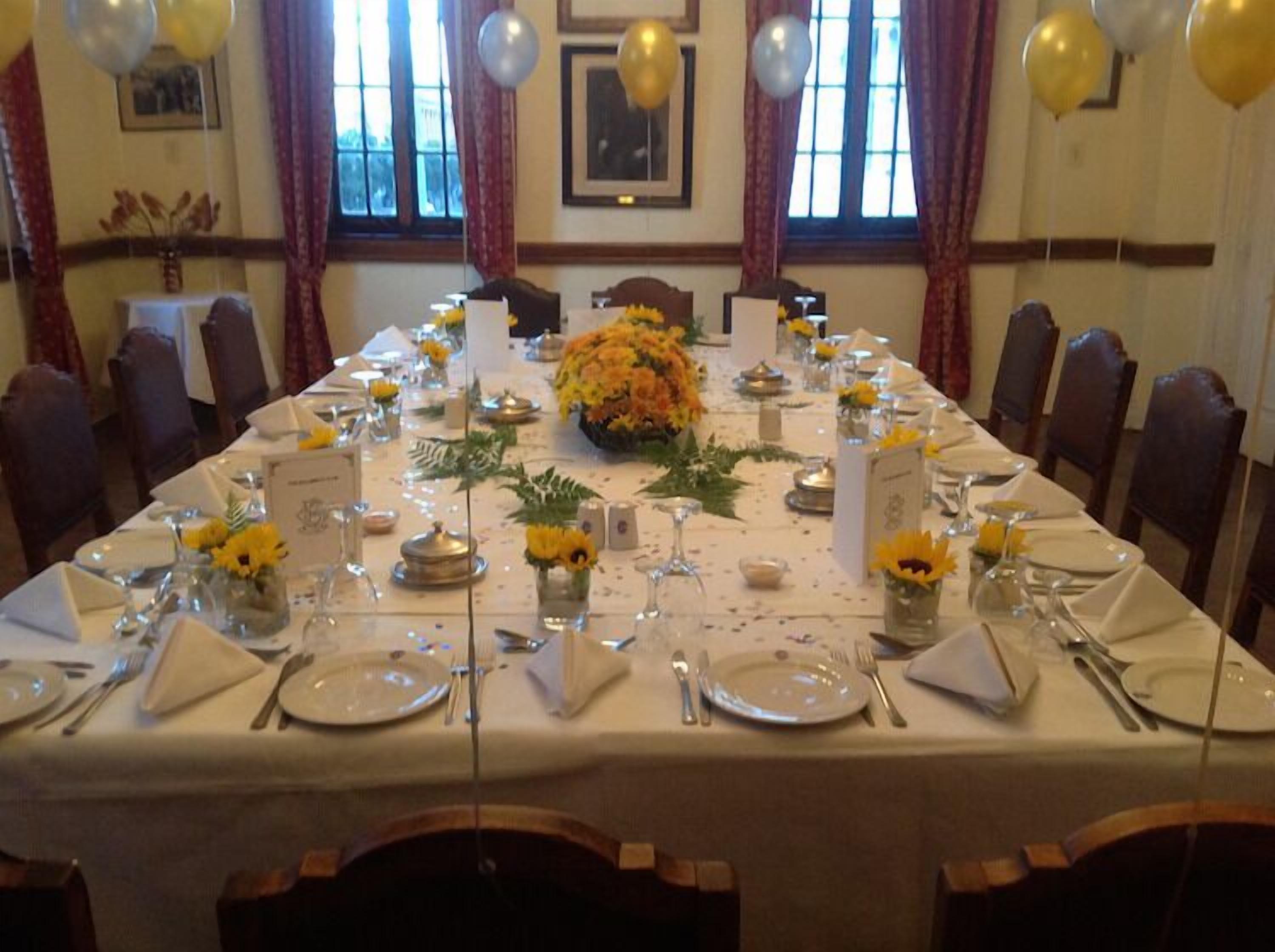 A long dining table set for a formal event with floral centerpieces, white tablecloths, and gold balloons.