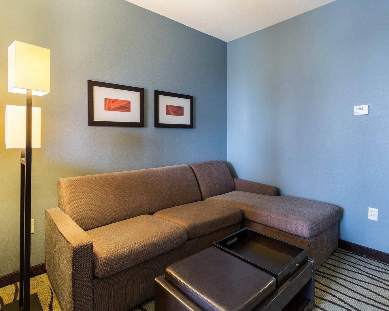A room with a sectional sofa, a small table, a floor lamp, and two framed pictures on the wall.