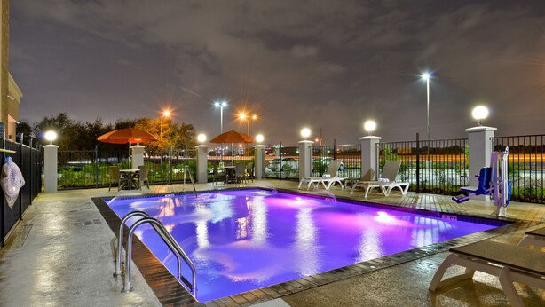 An outdoor swimming pool with a metal ladder, surrounded by lounge chairs and umbrellas, under a night sky with streetlights. - Comfort Suites near Westchase on Beltway 8 (Houston)