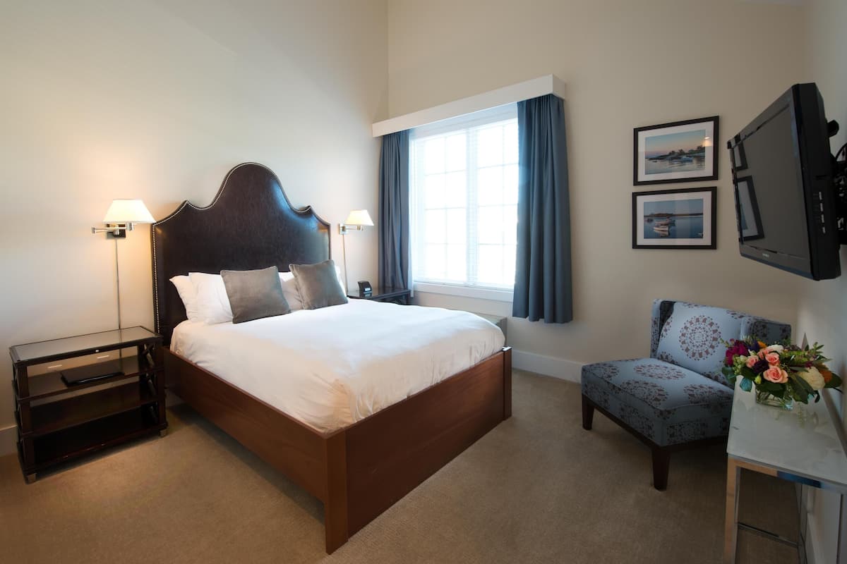 A hotel room with a large bed, bedside lamps, a desk with a monitor, a chair, and a window with curtains.