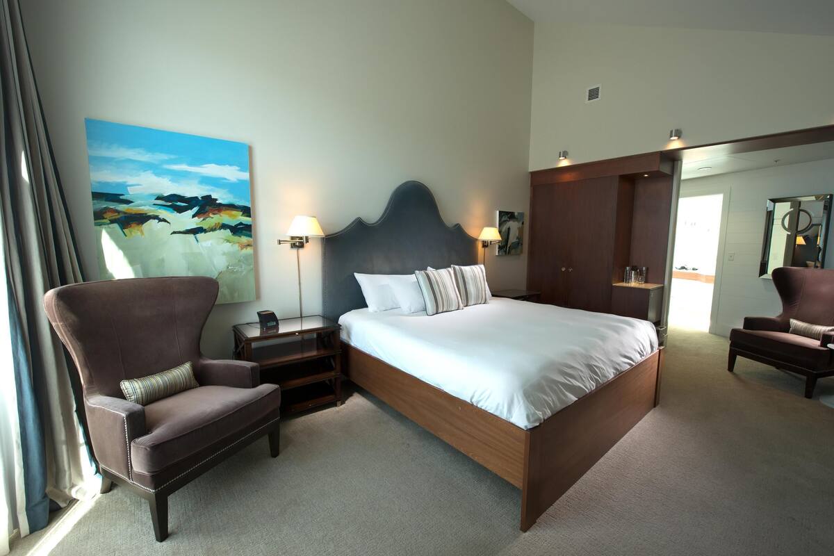 A modern hotel room with a large bed, a seating area with a sofa and armchair, a painting on the wall, and a wardrobe.