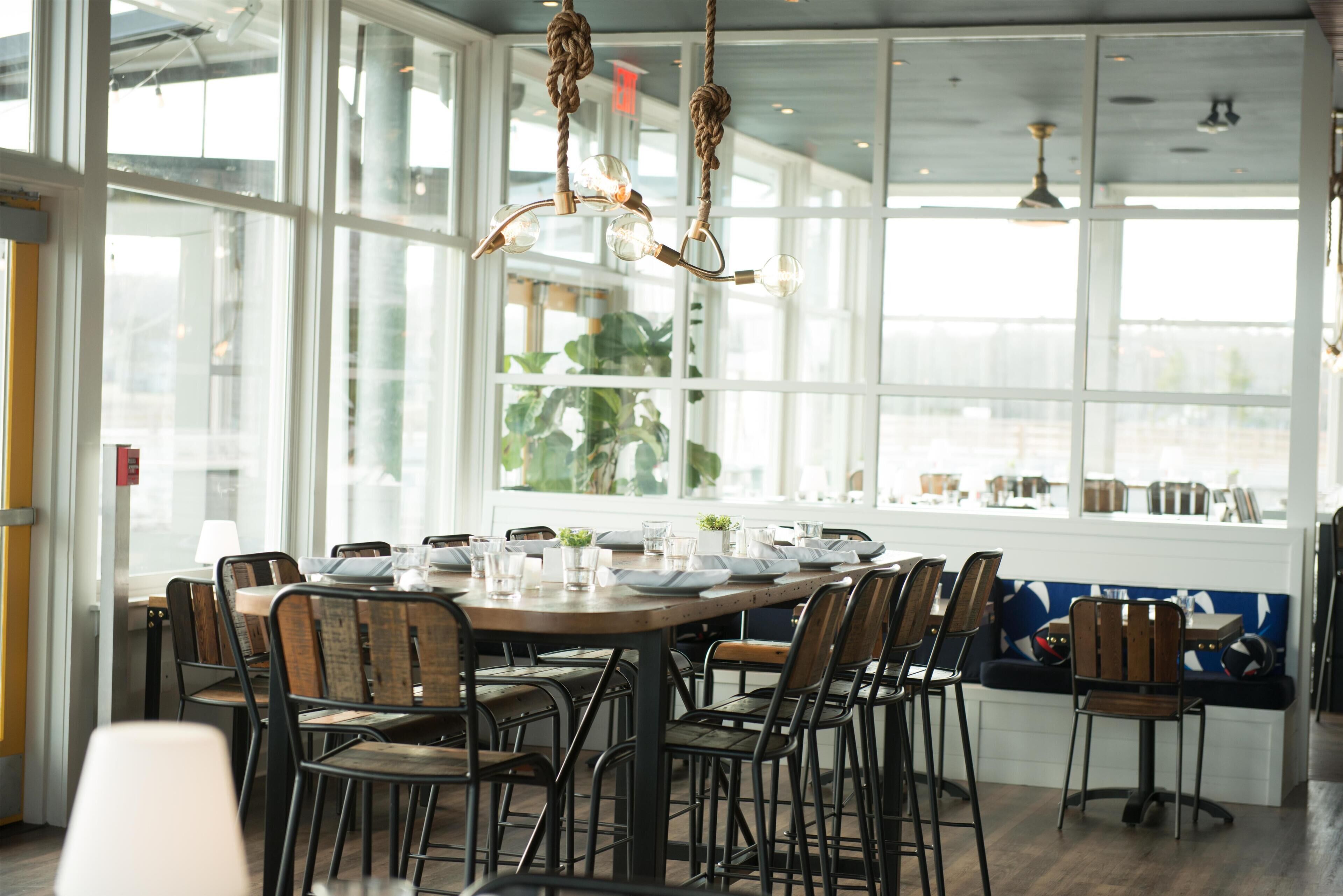Un espace repas avec une longue table dressée pour accueillir plusieurs personnes, entourée de chaises, de grandes fenêtres et une vue sur l’extérieur.
