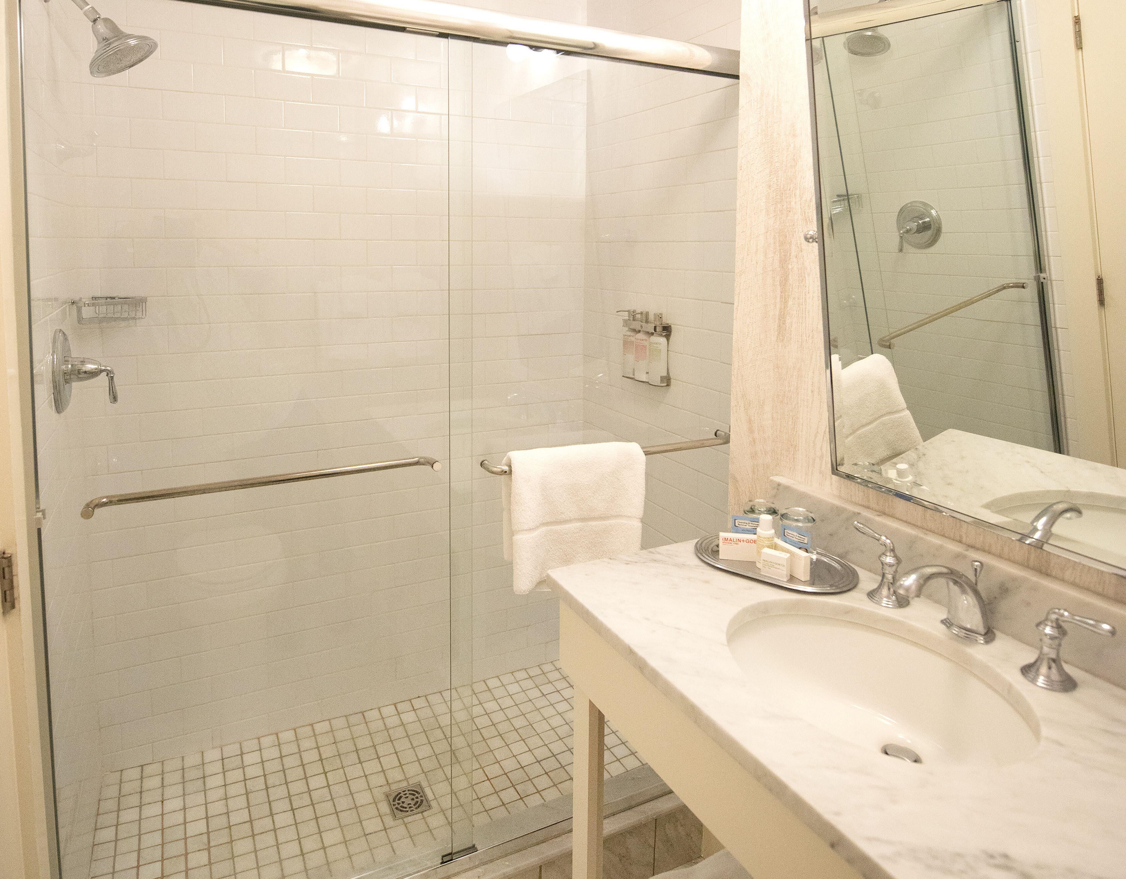 Un baño con ducha envuelta en vidrio, lavamanos con encimera de mármol y espejo sobre el lavamanos.