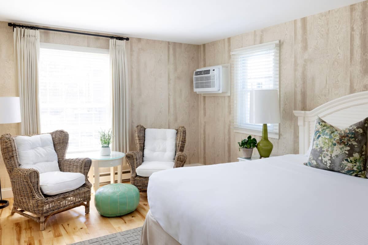 A hotel room with a large bed, two wicker chairs, a small round ottoman, a round table, a lamp, and a potted plant.