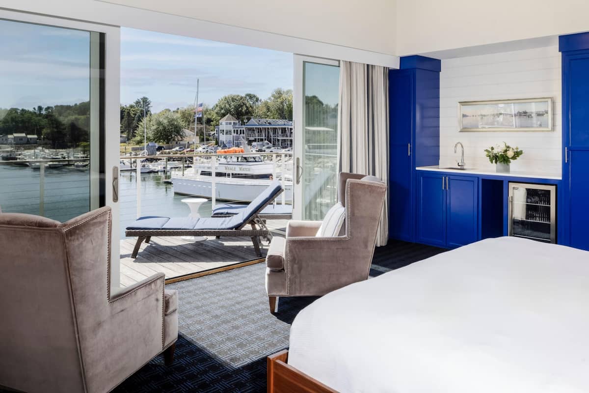 A hotel room with a large bed, two armchairs, a small table, a view of a marina with boats, and a blue accent wall.