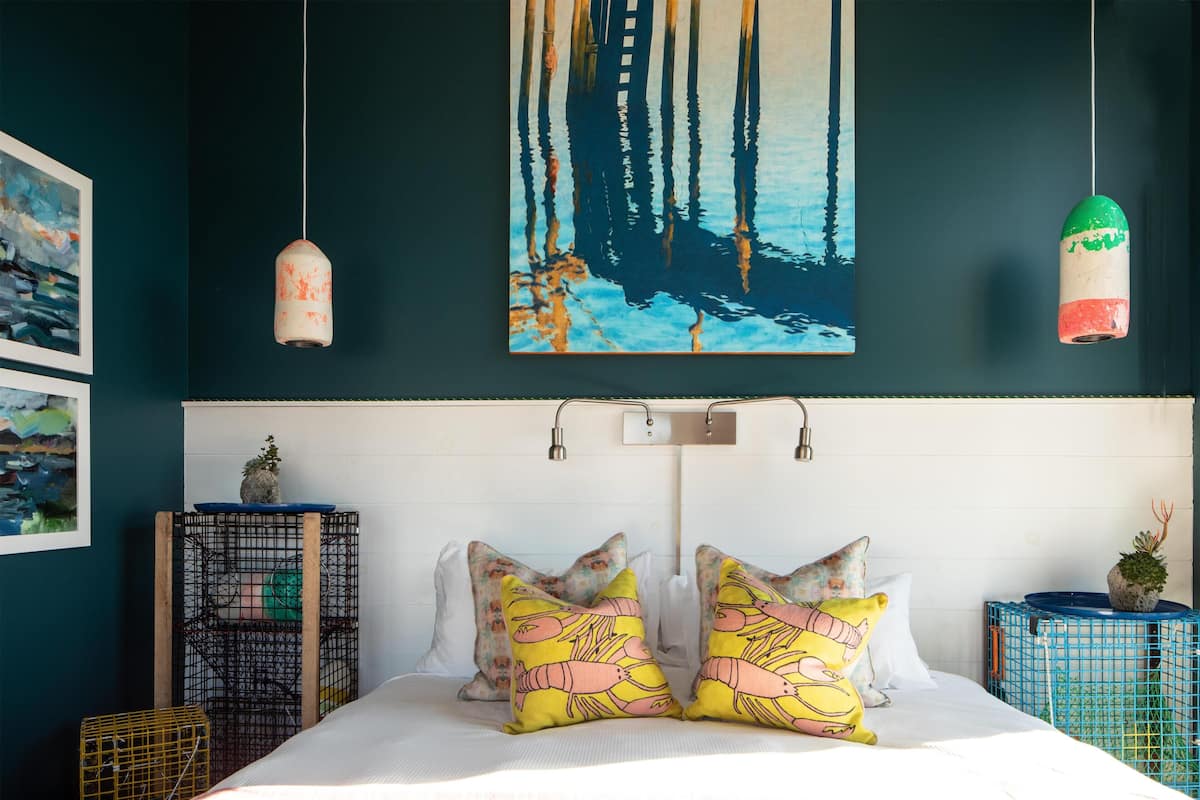 A bedroom with a bed, pillows, a painting, and a hanging light fixture.