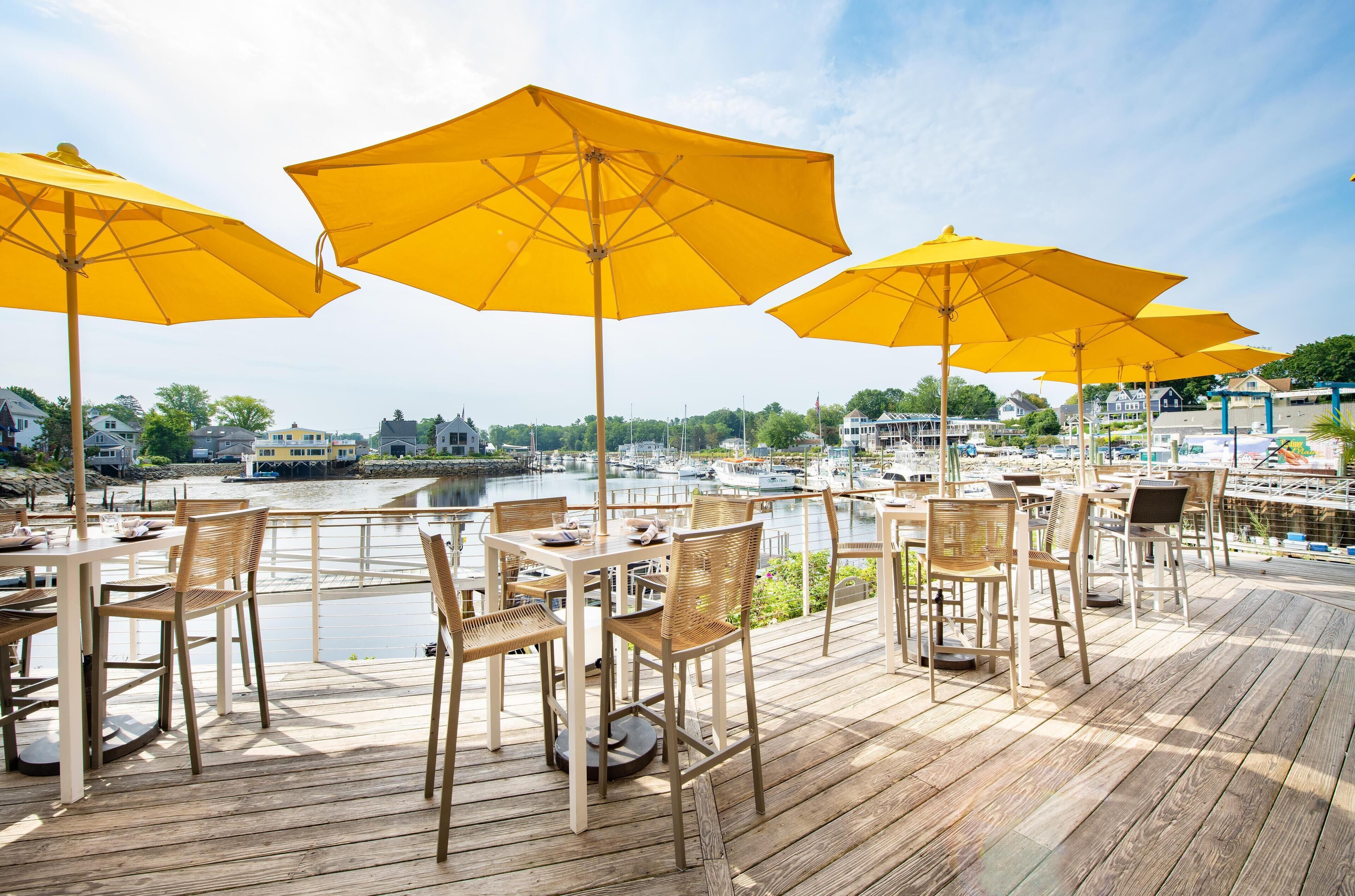 Eine Terrasse mit Holztischen und -stühlen, gelben Sonnenschirmen und Blick auf einen Yachthafen mit Booten.