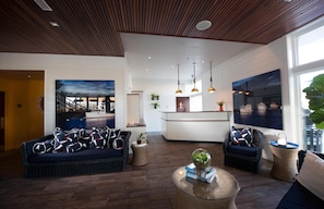 A modern hotel lobby with a dark wood ceiling, a large painting, a reception desk, and two seating areas with wicker furniture.