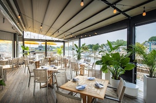 Un área de comedor al aire libre con mesas y sillas de madera, plantas en macetas y vistas de la ciudad.
