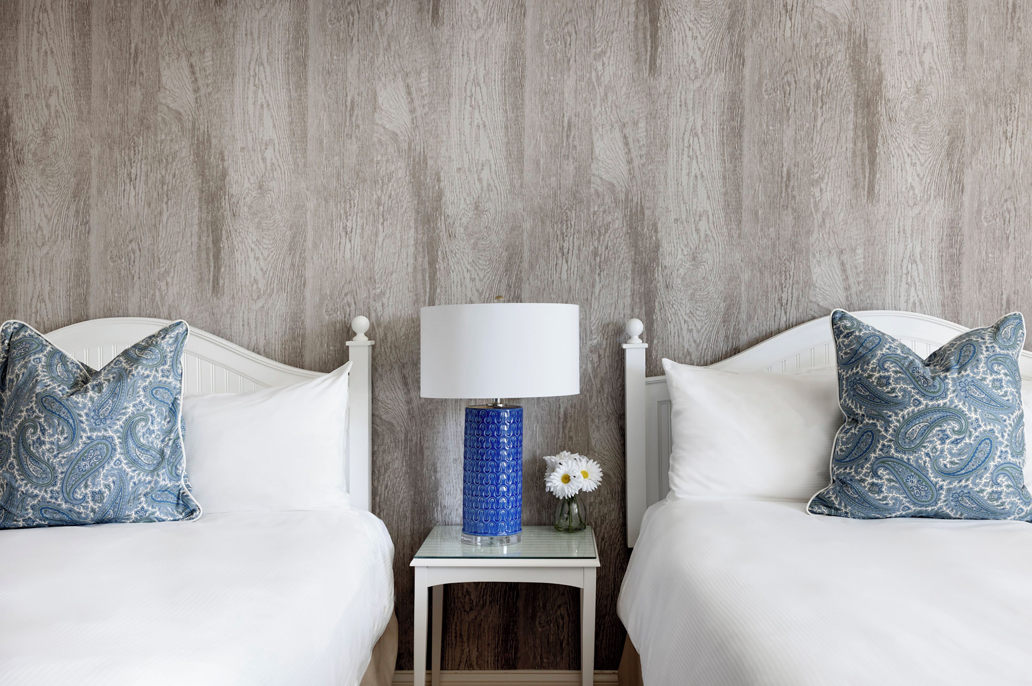 A hotel room with two beds, a small bedside table with a lamp, and a vase with a flower.