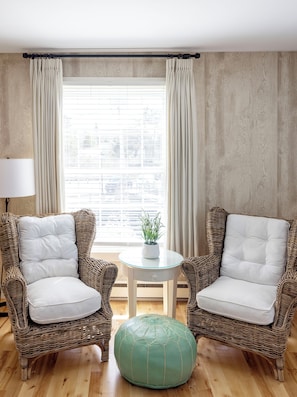 A room with two wicker chairs, a small round ottoman, a table with a plant, and a window with curtains.